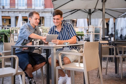 Gay couple sitting at an outdoor cafe in Plaza Mayor, Madrid, smiling while using a tablet and
