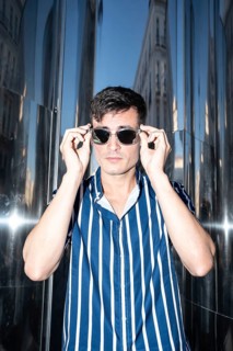 Fashionable young man in a striped shirt adjusting his sunglasses while posing against a reflective