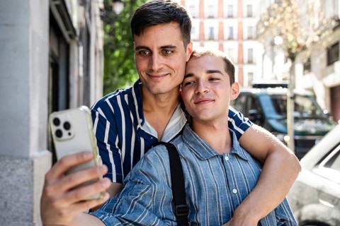 A joyful gay couple takes a selfie while embracing on the lively streets of Madrid. Their