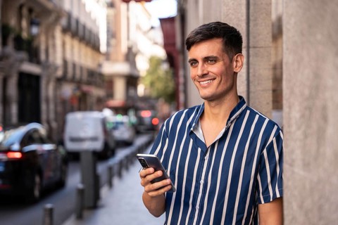 Stylish young man holding a smartphone and smiling on a vibrant city street. Dressed in a striped