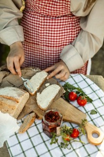 A person wearing a red checkered apron spreads cream on bread slices, surrounded by fresh tomatoes