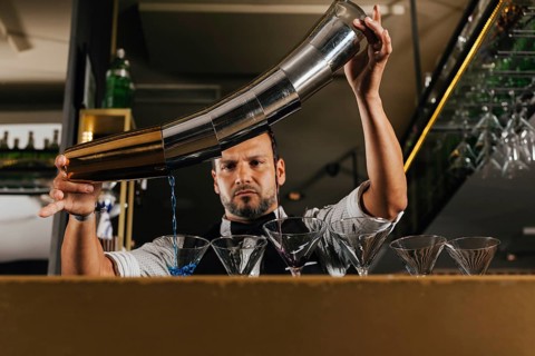 A professional bartender elegantly pours a vibrant blue cocktail into lined-up glasses at a chic