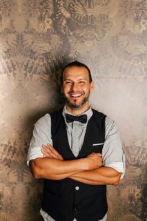 A professional bartender stands confidently with folded arms, wearing a stylish vest and bow tie.