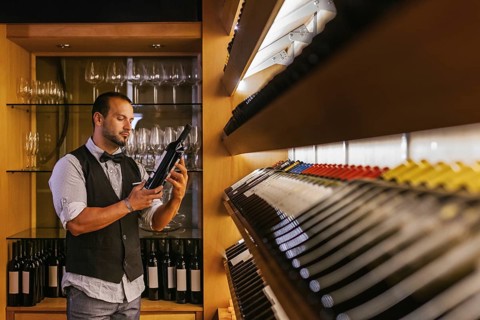 A professional sommelier selects a wine bottle in a stylish cocktail bar. Glasses line the shelves,