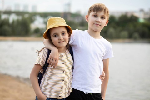 A young brother and sister share a warm moment by a lake. The boy, in a white shirt, lovingly puts