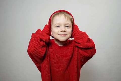A cheerful young child wearing an oversized red sweater and hat, smiling playfully at the camera.