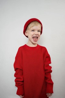 A cheerful child in a cozy red sweater and matching beanie laughs joyfully against a plain backdrop