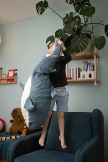A child joyfully playing with a large shark plush toy while standing on a chair in a cozy, well