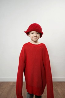 A child wears an oversized red sweater and matching hat, smiling playfully. The neutral background