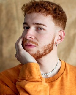 A close up portrait of a stylish young man with red curly hair, wearing a pearl necklace and