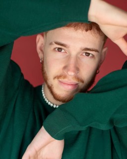 A man red hair wearing a green sweater poses with arms creatively framing their face against a red