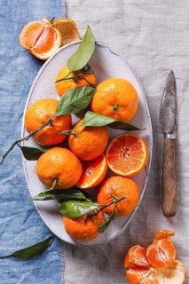 A bowl of vibrant tangerines with green leaves on a rustic table, accompanied by a vintage knife