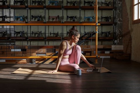 Woman in athletic attire practices yoga on a mat while working on a laptop. Sunlight filters