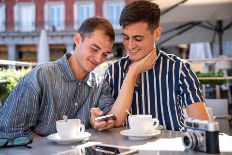 A gay couple shares a joyful moment at an outdoor cafe in Madrid. They smile while looking at a