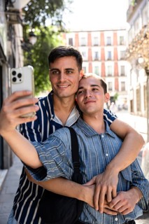 A gay couple embraces while taking a selfie on a picturesque street in Madrid. Sunlight filters