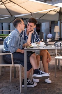A gay couple shares a joyful moment over coffee at an outdoor cafe in the vibrant streets of