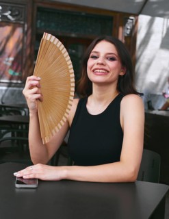 A joyful woman in a black tank top holds a hand fan, sitting at an outdoor cafe table with a