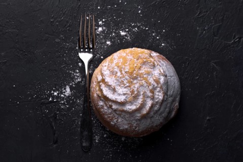 Top view of a single cream bun, generously sprinkled with icing sugar, rests next to a fork on a