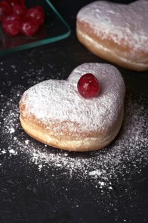 Two heart shaped cream buns, generously dusted with icing sugar and topped with a bright red