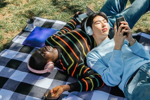Two friends enjoy a sunny day, lying on a checkered blanket. One listens to music with headphones,
