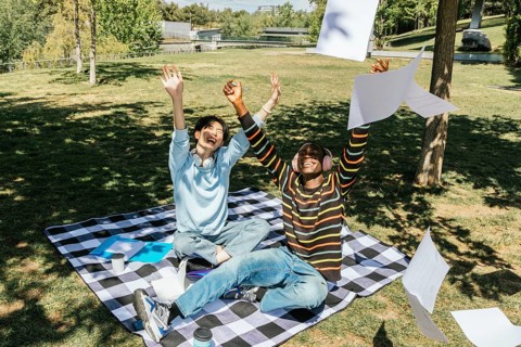 Two university students celebrate joyfully in a sunny park. Sitting on a picnic blanket, they toss