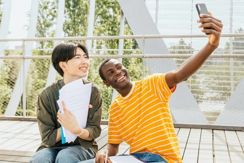 Two friends sit outdoors, smiling while taking a selfie. One holds papers, suggesting a study