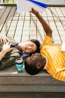 Two friends lying on a wooden deck. One is holding papers above them, while a coffee sits nearby.