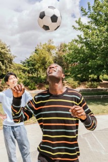 Two friends enjoy a sunny day in the park, playing soccer and laughing. The man in the foreground
