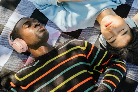 Two friends lie on a picnic blanket, enjoying music on headphones under the sunlight. Peaceful