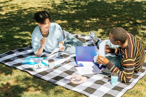 Two friends enjoy a study session outdoors on a sunny day, sitting on a checkered blanket with
