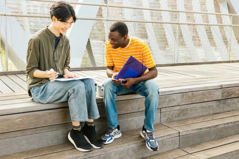 Two young adults study together outside on wooden steps. They are smiling and sharing notes. The
