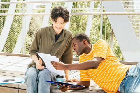 Two friends sit on wooden steps outdoors, discussing documents. One, in a striped shirt, leans in