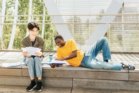 Two friends study outdoors on a sunny day, sitting and laying on a wooden steps surrounded by