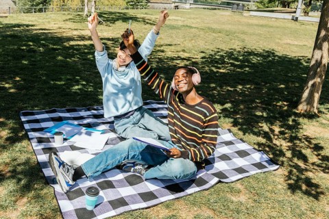 Two students friends are sitting on a checkered blanket in a sunny park. They are celebrating and