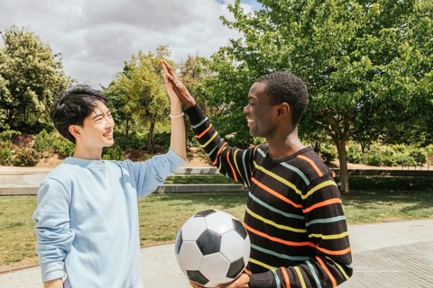 Two friends share a high five in a sunny park, celebrating after a soccer game. One holds a soccer