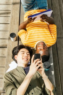 Two friends enjoy a sunny day outdoors. One is lying back with a notebook, while the other smiles