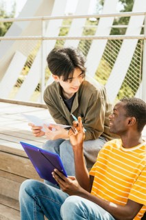 Two friends sit outside on wooden steps, engaged in a study session. One holds a blue folder, and
