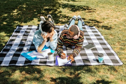 Two friends lie on a checked picnic blanket in a sunny park, sharing headphones, documents, and