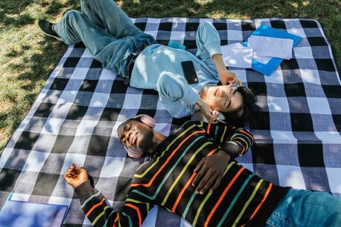 Two friends enjoy a sunny day outdoors, lying on a plaid blanket and listening to music with