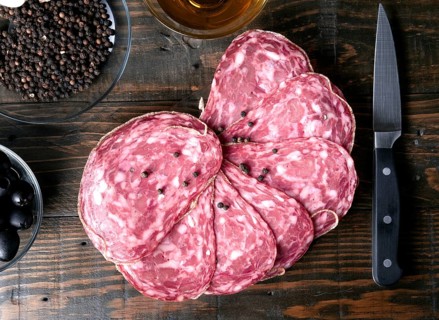 A top down view of sliced spanish salami, peppercorns, black olives, and a knife on a rustic wooden