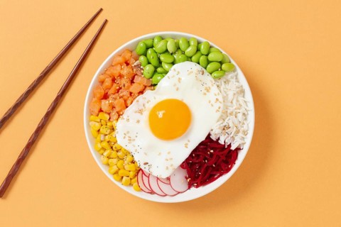 A colorful poke bowl featuring fresh ingredients like edamame, corn, salmon, beets, and a sunny