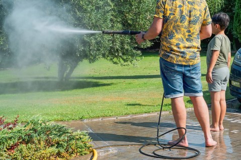 A father and child clean the garden with a pressure washer in summer setting, surrounded by trees
