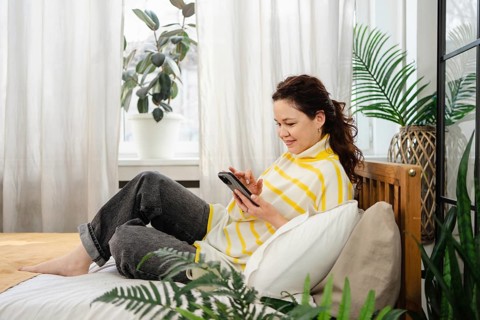 A woman sits comfortably on a bed, wearing a striped top, using her phone. The room is filled with