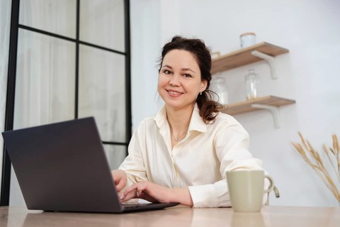 A professional woman, smiling and working on her laptop at a home office desk. Shelves and plants