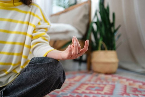 A woman meditates indoors, seated on a colorful rug. Their fingers are poised, surrounded by cozy,