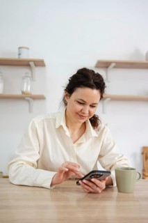 A woman in a cream shirt sits at a wooden table at home using her smartphone, next to a green