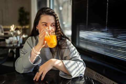 A mid-aged woman sips an orange drink in a brightly lit cafe, her expression serene and content as