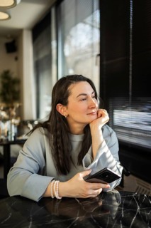 A middle-aged woman sits peacefully in a cafe, gazing outside while holding a smartphone in her