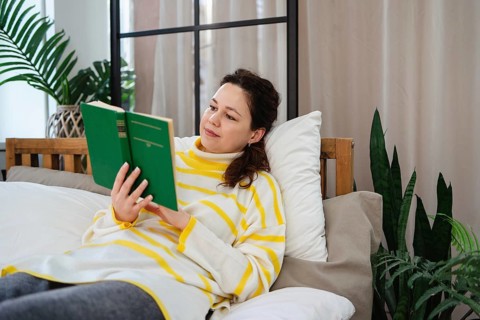 A woman wearing a striped sweater lies on a comfortable bed, deeply engrossed in reading a green