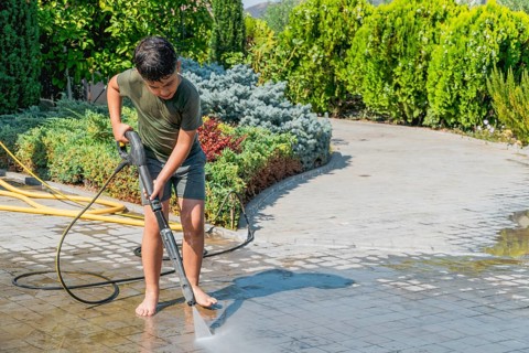 A child is using a pressure washer to clean a stone patio for the summer. He is barefoot, wearing a